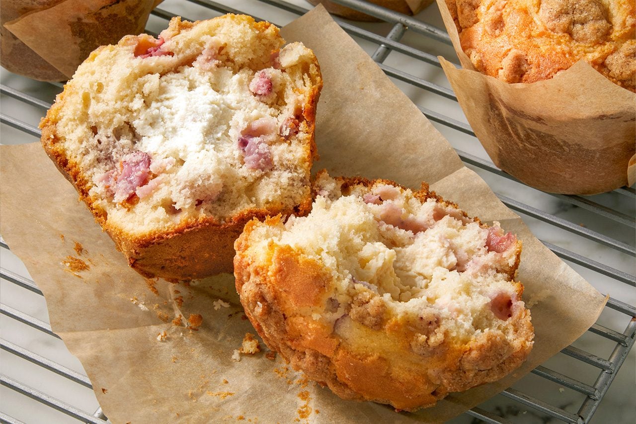 Eye Level horizontal image of a freshly baked muffin, broken in half, revealing its creamy white filling and chunks of fruit. Another whole muffin in a brown paper liner is visible in the background, all resting on a wire cooling rack.
