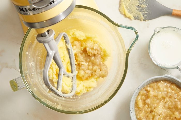 A stand mixer with creamed batter and mashed bananas in a glass bowl, next to a bowl of more mashed bananas and a small pitcher of milk on a light countertop.