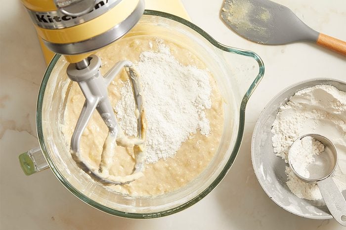 A stand mixer with a glass bowl is mixing batter and flour. Next to it is a bowl of flour with a metal measuring cup and a gray spatula on the counter.