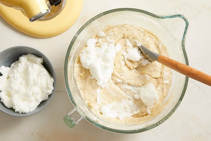A glass mixing bowl with batter and fluffy egg whites being folded in, a rubber spatula resting inside. Nearby, a small gray bowl holds more whipped egg whites, and part of a yellow stand mixer is visible.
