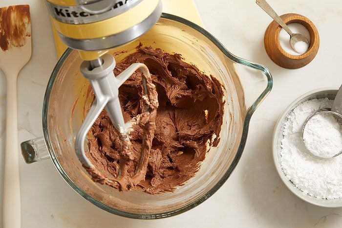 A stand mixer bowl with chocolate batter, a mixing paddle attached, sits on a counter near powdered sugar in a bowl, a small wooden container with a spoon, and a spatula.