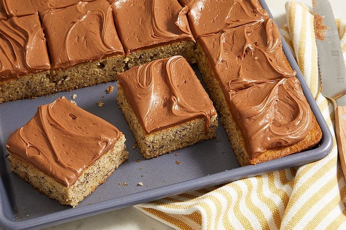 A baking pan with square slices of cake topped with chocolate frosting sits on a striped cloth. A serving spatula rests nearby.