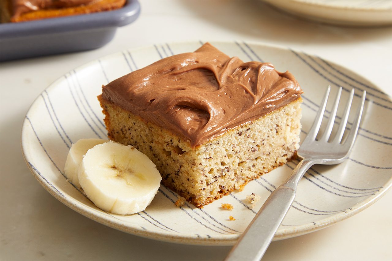 A slice of banana cake with chocolate frosting is on a striped plate, next to two banana slices and a fork.