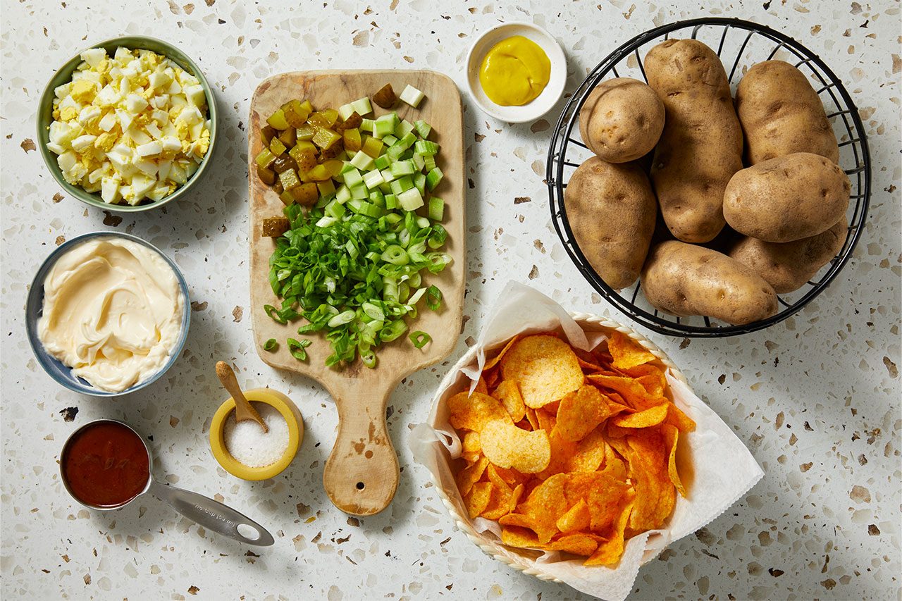 Overhead horizontal image of various ingredients prepared on a speckled white countertop. Chopped hard-boiled eggs, mayonnaise, chopped pickles, celery, and green onions are neatly arranged, alongside whole potatoes, yellow mustard, salt, a dark sauce in a measuring cup, and a basket of potato chips.