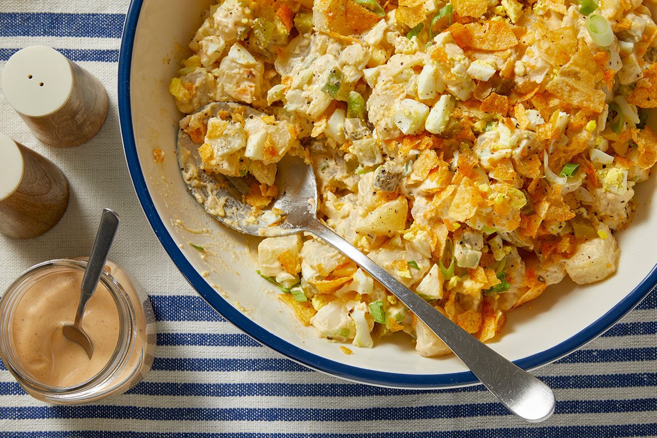 Overhead horizontal image of a large bowl of potato salad with a serving spoon, garnished with crushed potato chips and sliced green onions. Visible ingredients include diced potatoes, chopped hard-boiled eggs, and green chopped vegetables, possibly celery or pickles. To the side are wooden salt and pepper shakers and a jar of creamy dressing with a spoon, all resting on a blue and white striped tablecloth.
