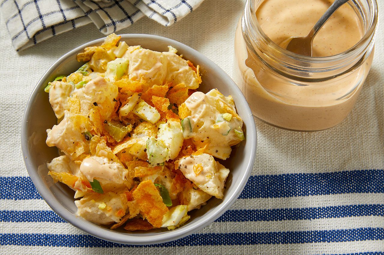Overhead horizontal image of a creamy salad in a gray bowl, featuring visible chunks of potato or egg, green bits like celery, and generously topped with golden crumbled chips. A glass jar with a spoon, holding additional creamy dressing, sits beside the bowl.