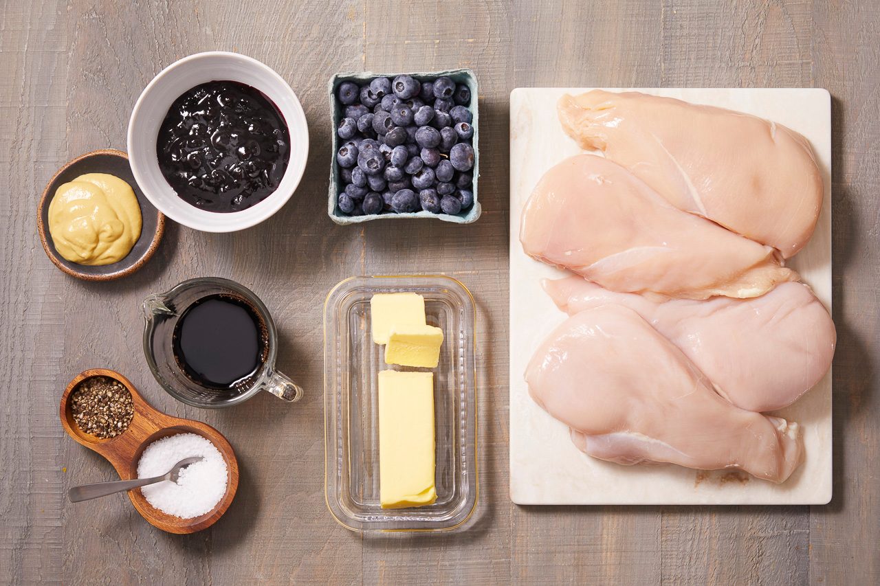 Overhead shot of raw chicken breasts on a cutting board alongside fresh blueberries, blueberry preserves, butter, Dijon mustard, balsamic vinegar, salt, black pepper, and seasonings arranged on a wooden surface.