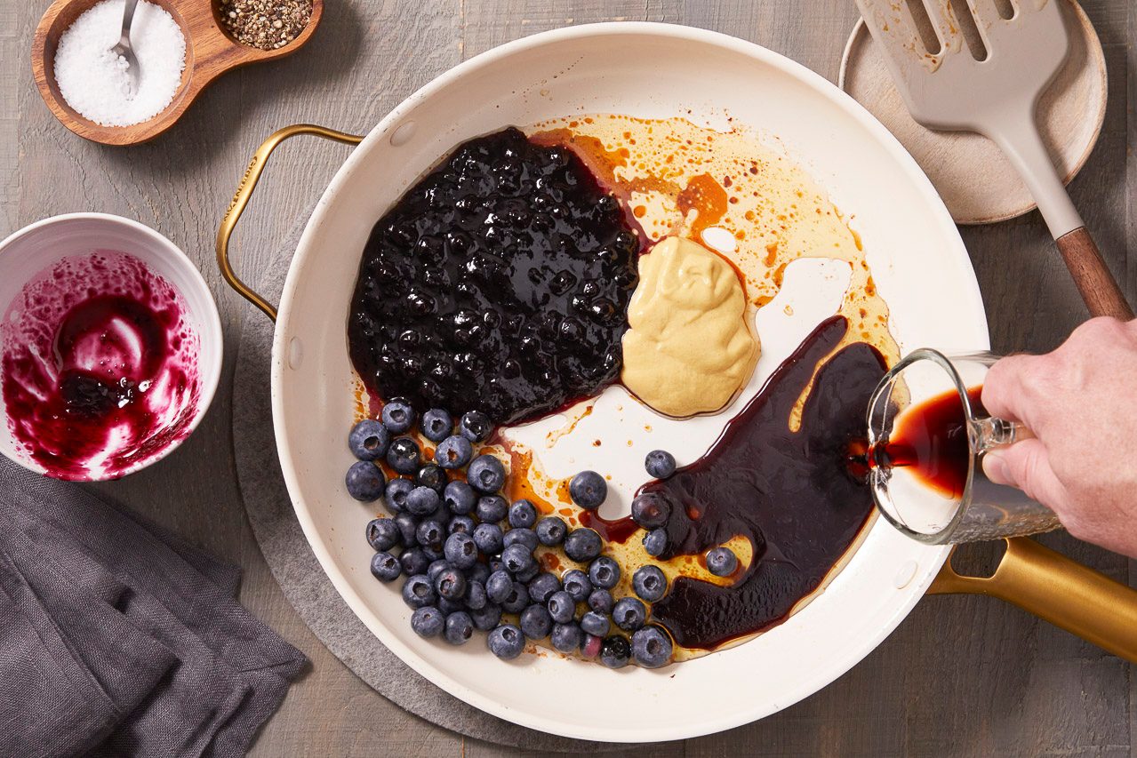Overhead shot of blueberry preserves, fresh blueberries, Dijon mustard, and balsamic vinegar being combined in a white skillet to prepare a fruit sauce.