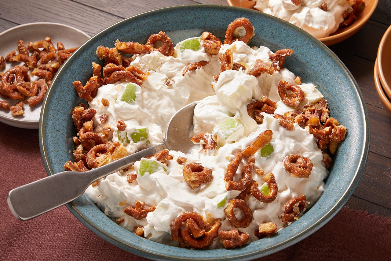 Eye Level horizontal image of a large blue bowl filled with a creamy dessert or salad. The white mixture is studded with green apple chunks and generously topped with candied pretzels and chopped pecans, with a silver serving spoon resting in the bowl.