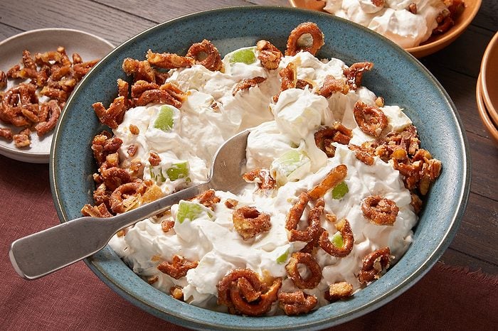 Eye Level horizontal image of a large blue bowl filled with a creamy dessert or salad. The white mixture is studded with green apple chunks and generously topped with candied pretzels and chopped pecans, with a silver serving spoon resting in the bowl.