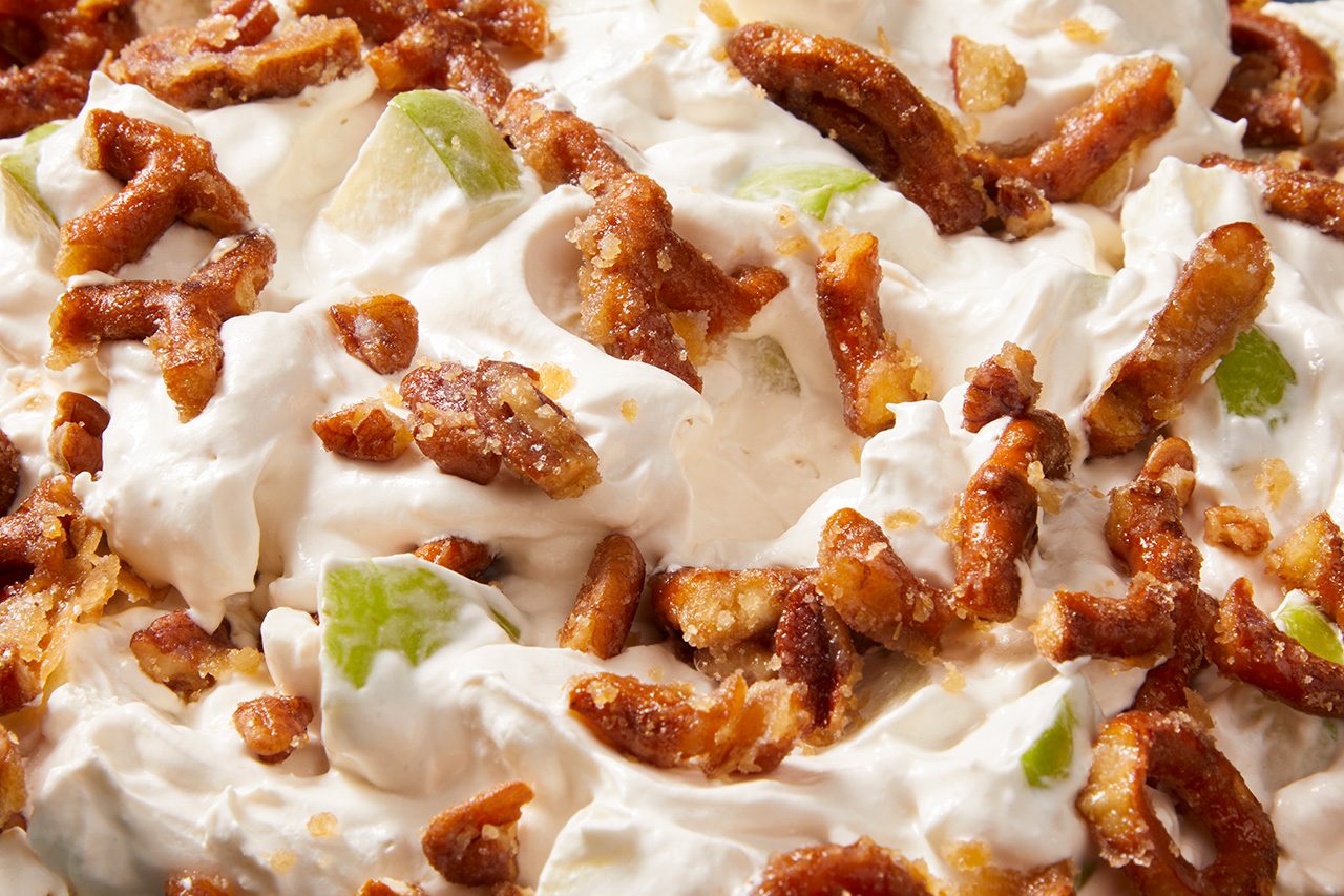 Overhead horizontal image of a creamy, white dessert base, likely whipped cream, mixed with crunchy candied pretzels. Small chunks of green apple are also visible throughout the sweet mixture.