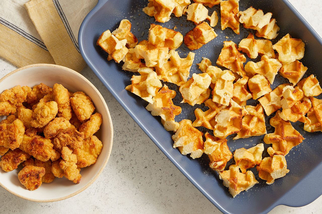 Overhead horizontal image of a bowl of fried chicken pieces next to a blue baking dish filled with cut-up waffle pieces. A striped yellow dishtowel is visible in the background on a light grey speckled surface.