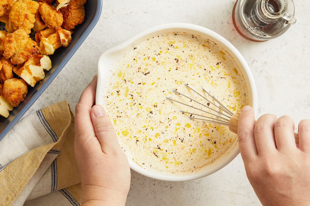 Overhead horizontal image of two hands whisking a creamy batter in a white bowl. The batter contains dark specks and yellow spots. A blue baking dish filled with fried chicken pieces and waffle cubes, along with a bottle of syrup, are also visible on the counter.