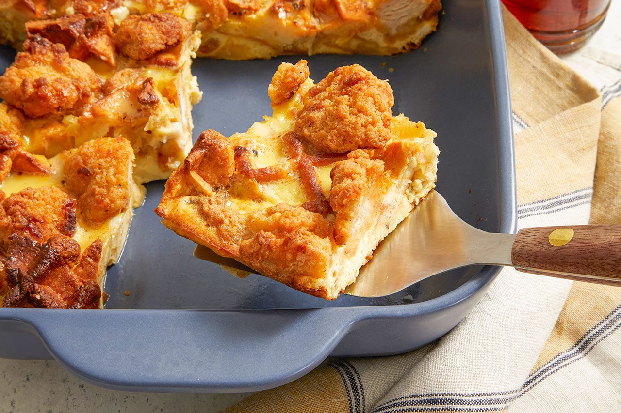 Overhead horizontal image of a serving of savory chicken casserole being lifted from a blue baking dish with a spatula. The casserole features golden-brown crispy fried chicken pieces and a creamy, baked egg base, with a striped linen napkin in the background.