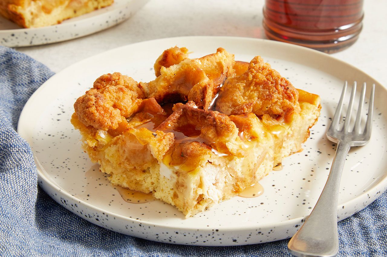 Eye Level horizontal image of a slice of bread pudding with crispy fried chicken pieces on top, drizzled with maple syrup. It rests on a speckled white plate next to a silver fork, with a blue napkin beneath it.