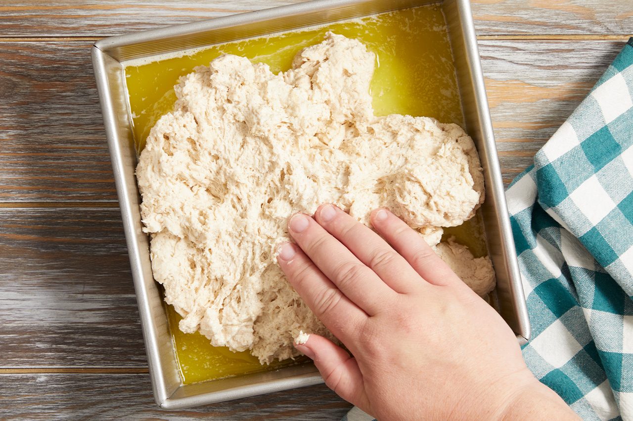 Overhead horizontal image of a hand pressing down on a lumpy dough mixture in a square metal baking pan. The pan contains a layer of melted butter at the bottom, partially submerging the dough. A blue and white checkered kitchen towel rests on a wooden surface next to the pan.