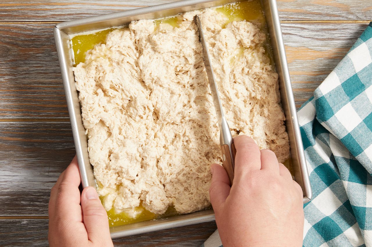 Overhead horizontal image of hands using a knife to score or cut dough in a square baking pan. Melted butter is visible at the bottom of the pan, and a teal and white plaid towel rests on a wooden surface beside it.