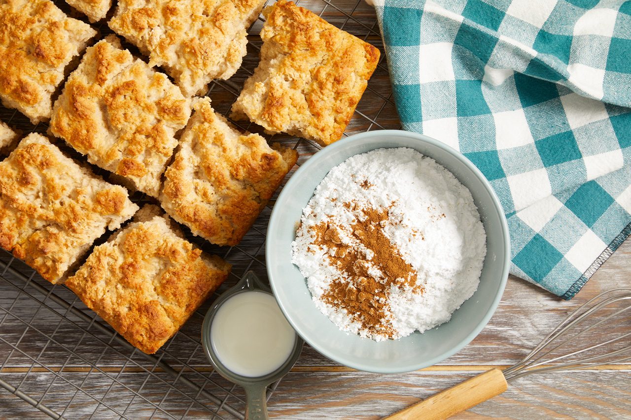 Overhead horizontal image of golden-brown square biscuits cooling on a wire rack. A light blue bowl holds powdered sugar and ground cinnamon, while a small measuring cup of milk and a whisk are placed nearby, suggesting preparation for a glaze or topping.