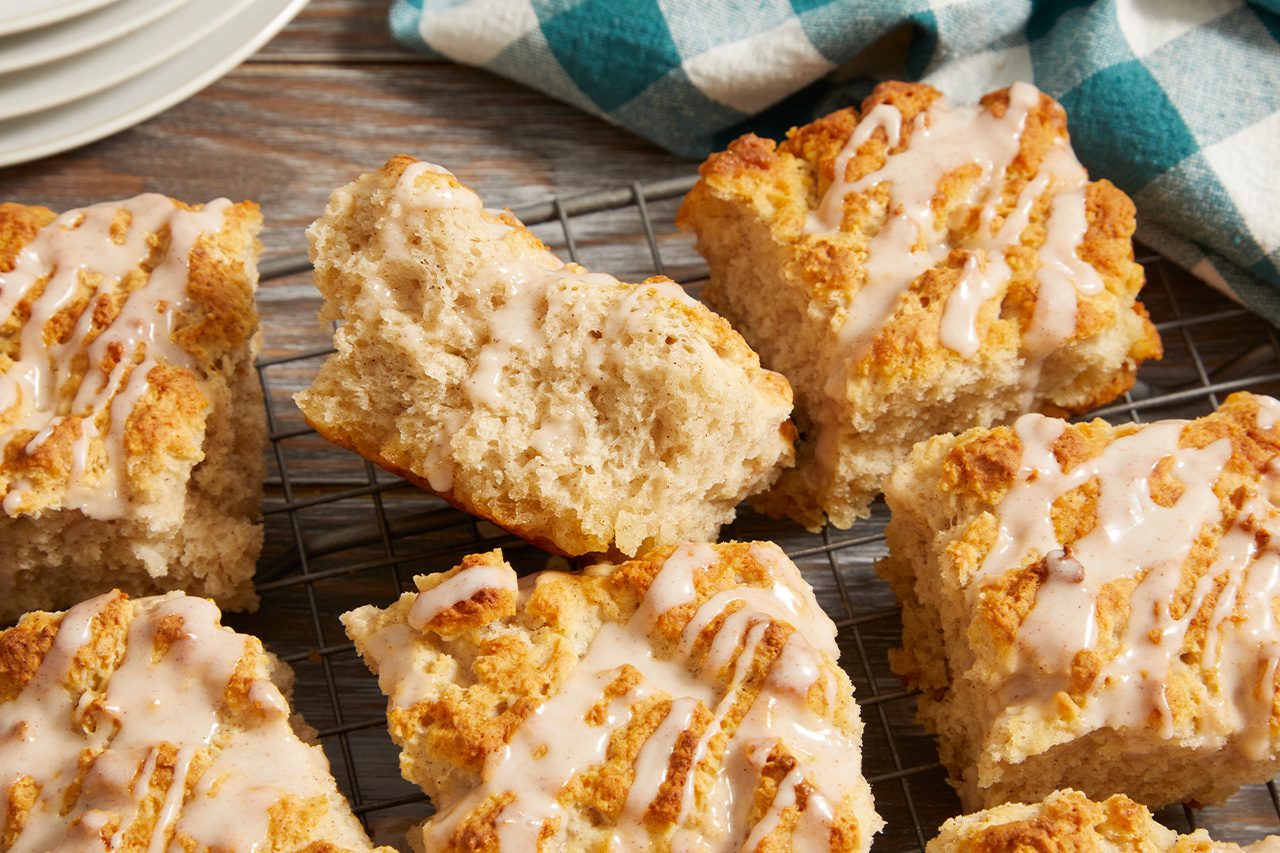 Overhead horizontal image of several square-shaped scones drizzled with a light brown icing, resting on a wire cooling rack. One scone is broken open, revealing its fluffy interior, with a checkered napkin and stacked plates visible in the soft-focus background.