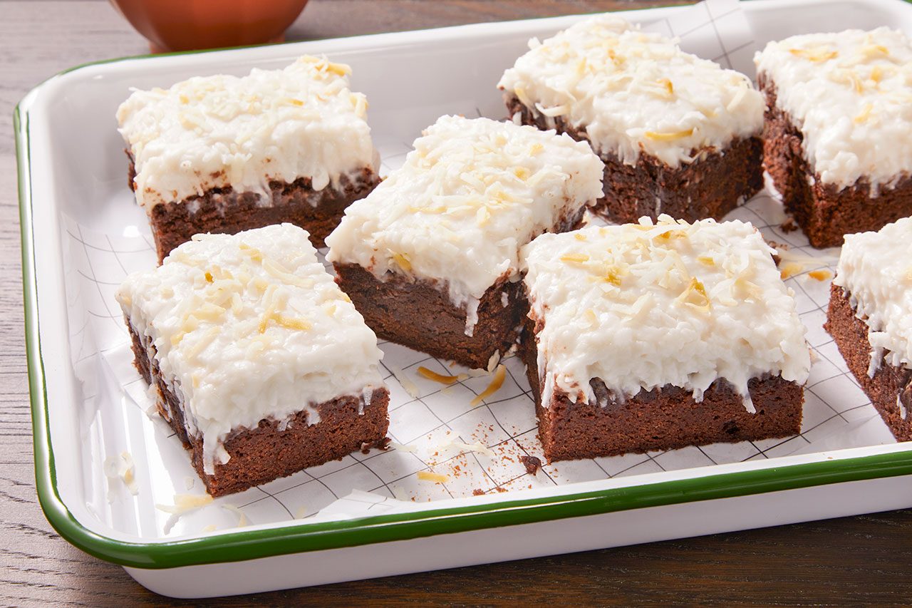 Eye Level horizontal image of several square-cut chocolate brownies in a white enamel baking dish. Each brownie is topped with a thick layer of white coconut frosting and garnished with toasted coconut flakes.