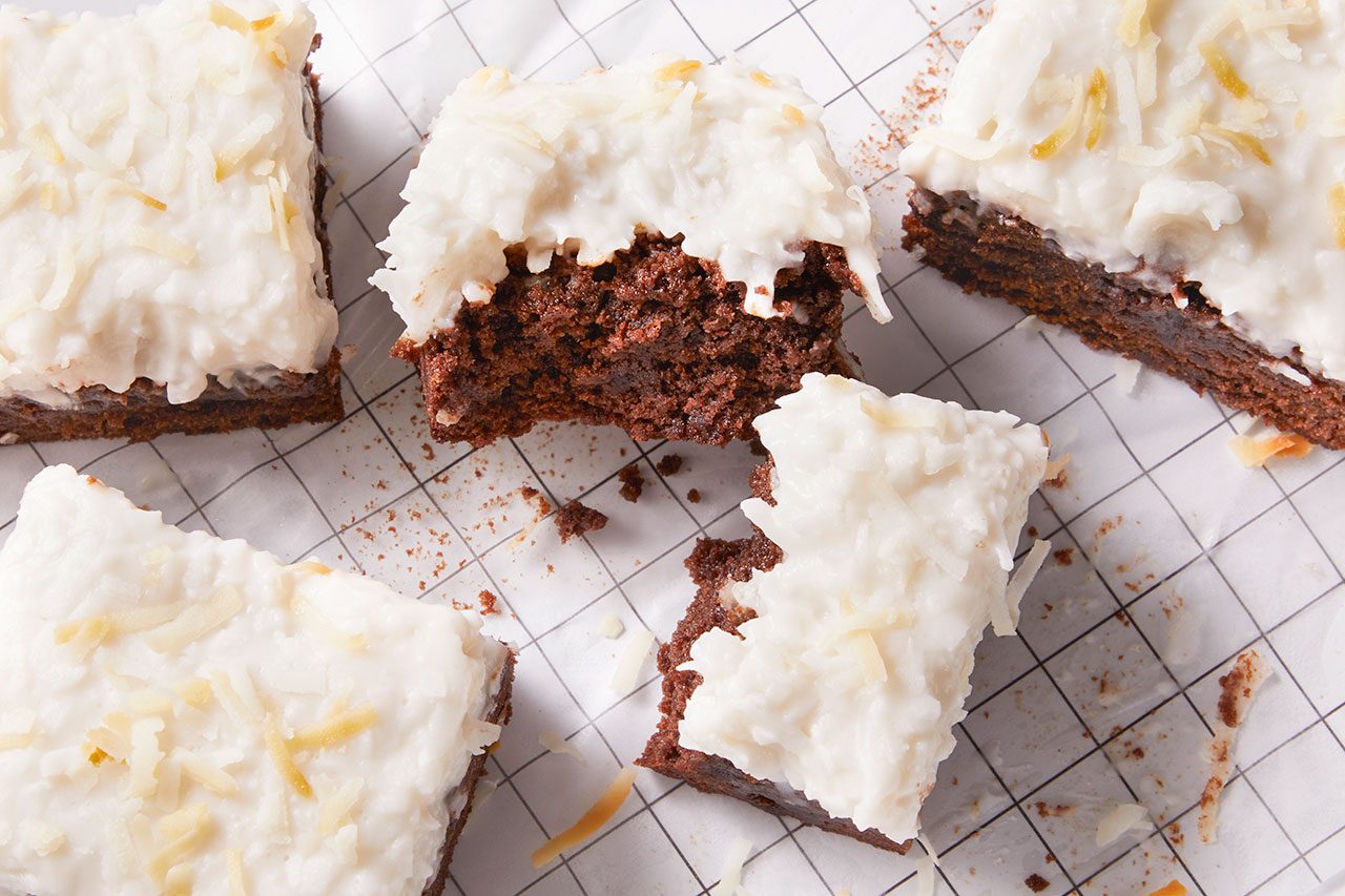 Overhead horizontal image of several square pieces of frosted brownies with shredded coconut on top, arranged on white paper with a grid pattern. One brownie has a bite taken out of it, revealing the rich chocolate interior. The coconut flakes are scattered across the white frosting, with some fallen onto the paper.