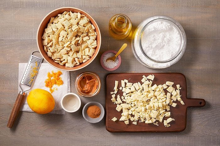 Overhead horizontal image of all ingredients for Cookie Butter Puppy Chow laid out on a wooden surface. A bowl of Rice Chex, chopped white baking chocolate, canola oil, confectioners' sugar, Biscoff creamy cookie spread, grated orange zest with an orange and grater, ground cinnamon, and kosher salt are ready for preparation.
