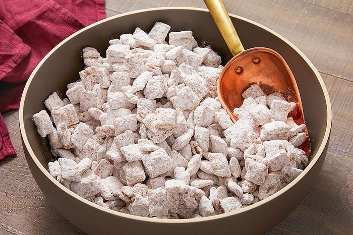 Eye Level horizontal image of a dark gray bowl filled with finished Cookie Butter Puppy Chow. The crispy cereal pieces are generously coated in white confectioners' sugar, with a copper scoop resting inside the bowl. A maroon linen napkin is visible in the background on a wooden surface.