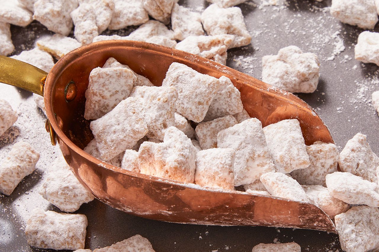 Overhead horizontal image of finished Cookie Butter Puppy Chow. The cereal pieces are heavily coated in confectioners' sugar, with many pieces filling a copper scoop and others scattered across a dark surface.