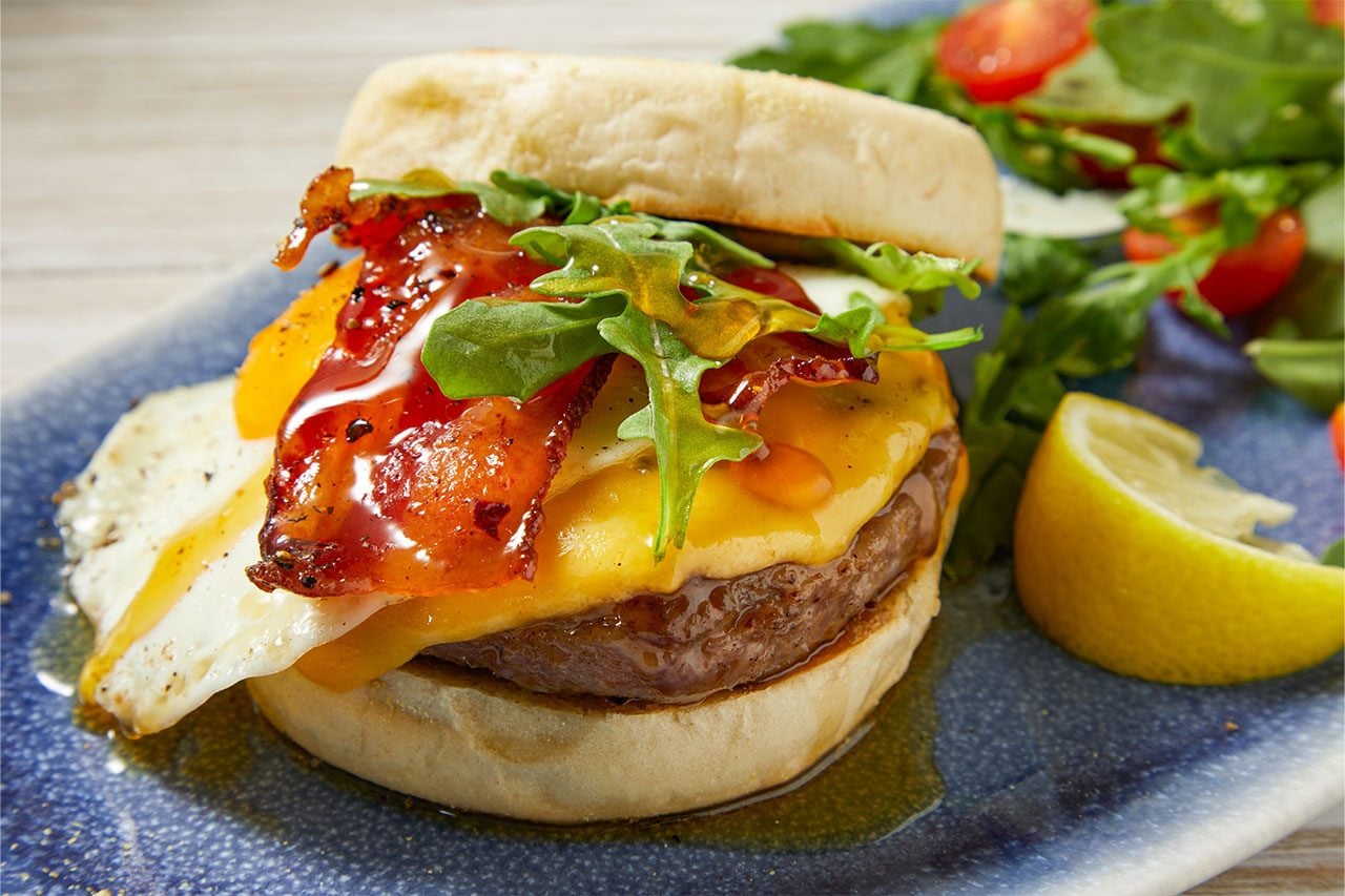 Close-up shot of Copycat First Watch Million-Dollar Breakfast Sandwich featuring a toasted English muffin layered with a sausage patty, fried egg, melted cheddar cheese, crispy bacon, and fresh arugula. The sandwich is served on a blue plate with a mixed green salad and a lemon wedge, highlighting its rich, hearty layers.