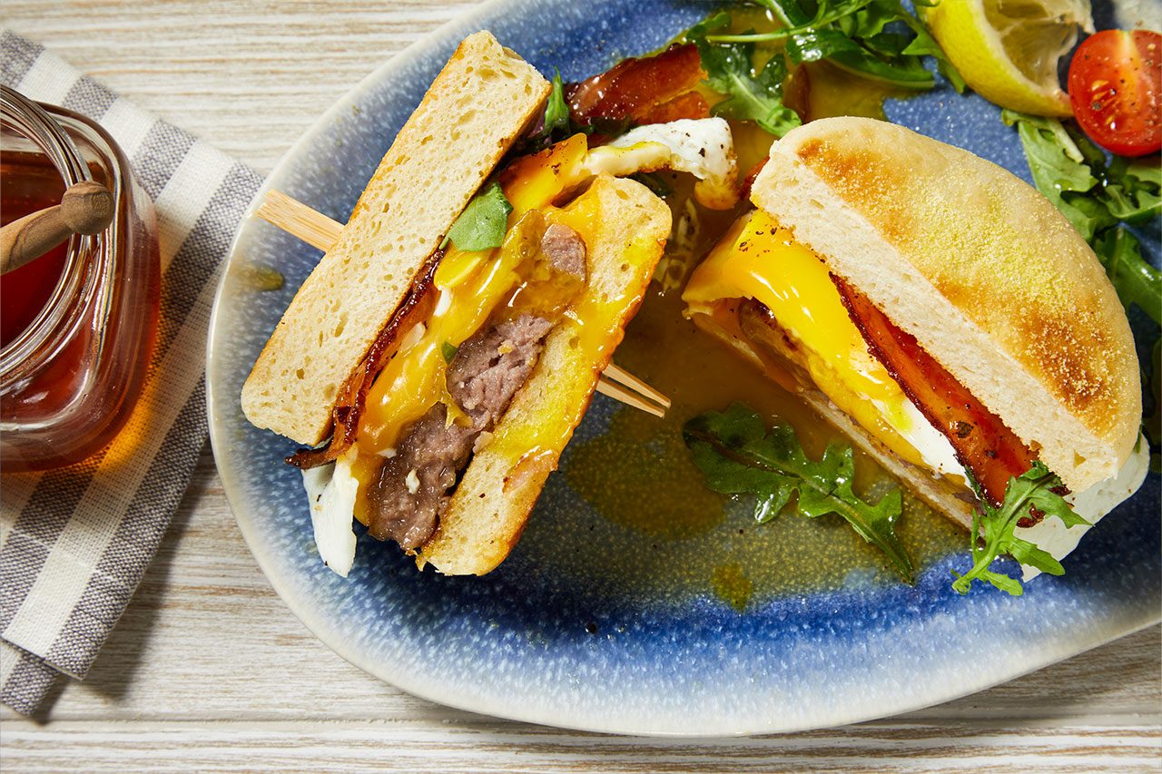 Close-up shot of Copycat First Watch Million-Dollar Breakfast Sandwich cut in half to reveal layers of sausage, fried egg, melted cheddar cheese, crispy bacon, and fresh arugula on a toasted English muffin. The sandwich is served on a blue plate with a mixed green salad, lemon wedge, and a jar of dressing nearby.