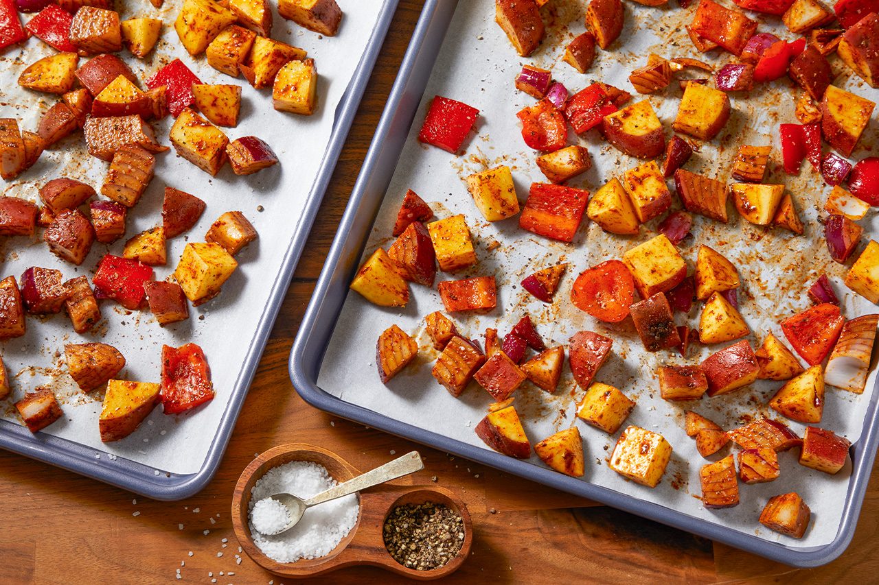 Overhead horizontal image of two baking sheets lined with parchment paper, filled with seasoned, chopped red potatoes, red bell pepper, and red onion. A small wooden dish containing coarse salt and ground black pepper is visible in the foreground on a wooden countertop.