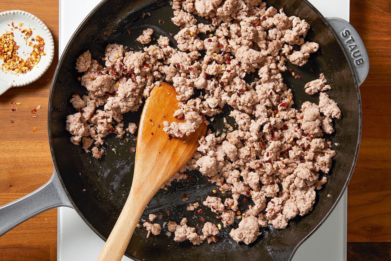 Overhead horizontal image of ground sausage browning in a black Staub skillet. A wooden spoon rests in the pan, stirring the meat which is mixed with red pepper flakes. A small dish of additional red pepper flakes is visible to the left of the skillet.