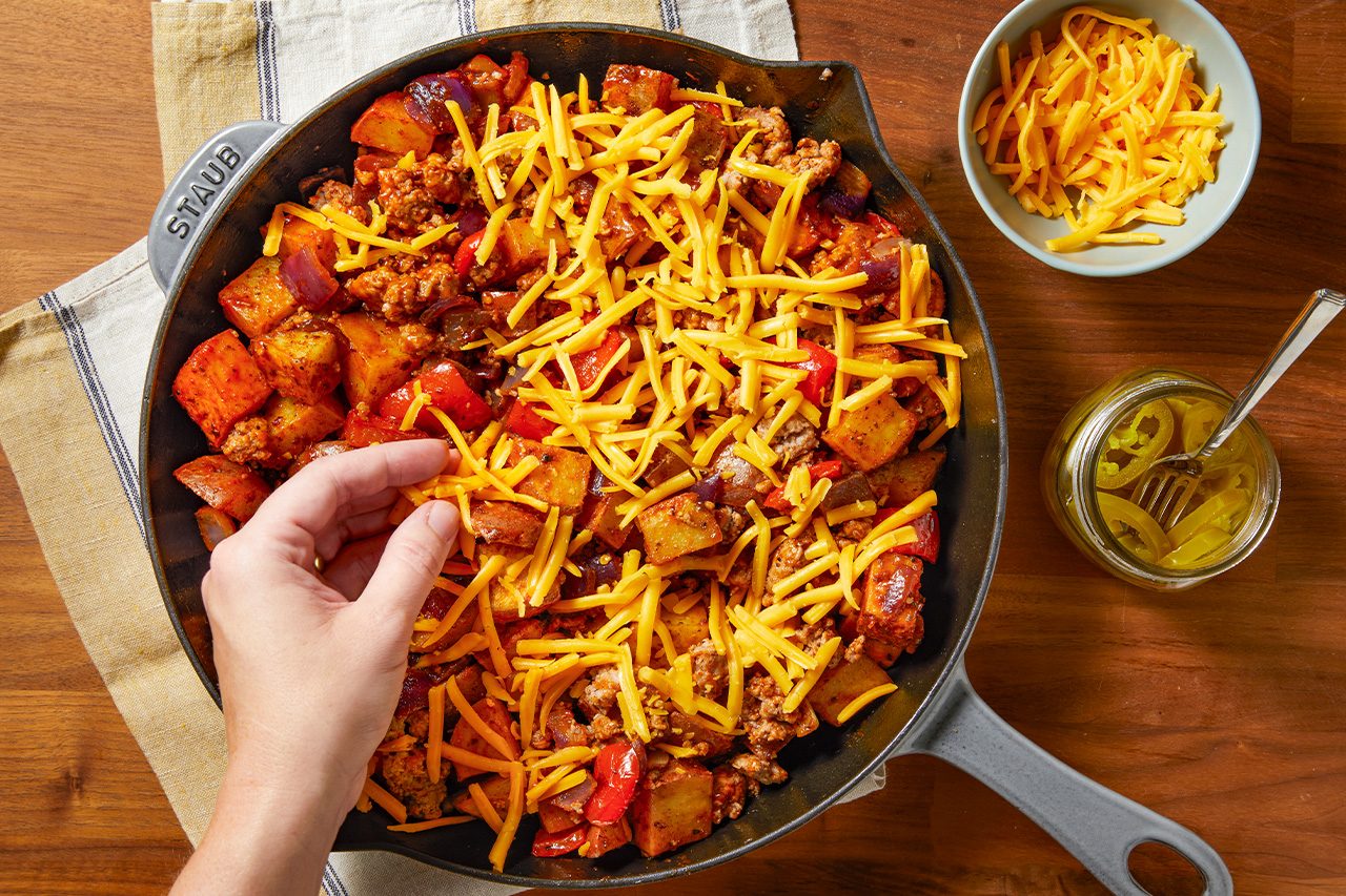 Overhead horizontal image of a hand sprinkling shredded cheddar cheese over a cast iron skillet filled with a savory mixture. The skillet contains ground meat, cubed potatoes, red bell peppers, and red onions. A bowl of extra shredded cheese and a jar of pickled jalapeños with a fork are visible to the side.