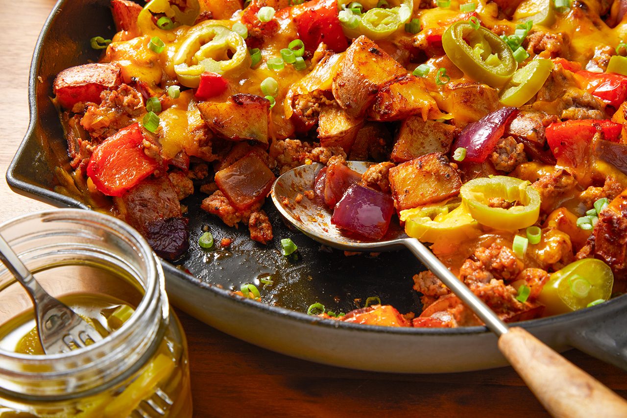 Eye-level horizontal image of a cast iron skillet filled with a savory cheesy potato and ground meat dish. The hearty meal, garnished with sliced pickled peppers and green onions, features roasted potatoes, ground meat, red bell peppers, and red onions. A serving spoon is in the skillet, with a jar of pickled peppers and a fork visible in the foreground.