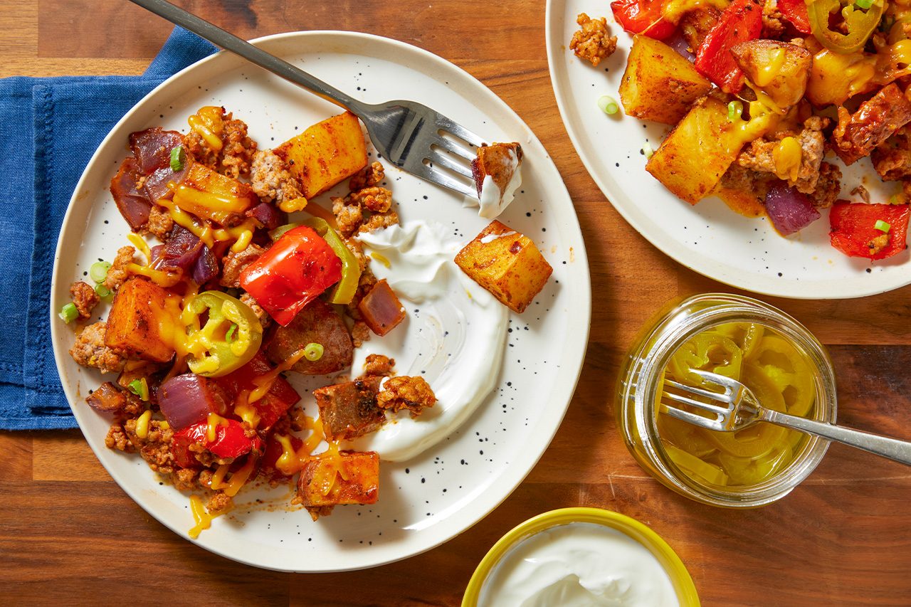 Overhead horizontal image of a white plate holding a serving of hearty hash made with cubed potatoes, seasoned ground meat, melted cheese, red bell peppers, red onions, and sliced jalapeños. A fork is spearing a potato chunk, partially dipped in a dollop of sour cream on the side of the plate. Another plate, a jar of pickled jalapeños with a fork, and a small yellow bowl of sour cream are also visible on the wooden table.
