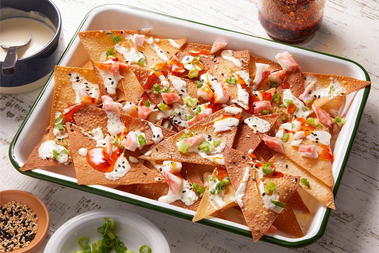Eye level horizontal image of an enamel baking dish filled with loaded wonton nachos. Crispy wonton chips are topped with chunks of imitation crab, a creamy white sauce, a vibrant chili sauce, chopped green onions, and a sprinkle of black and white sesame seeds.