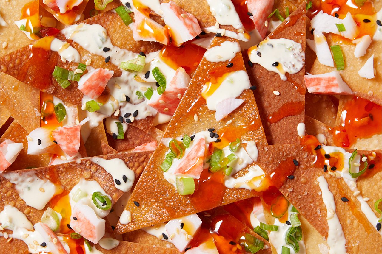 Overhead horizontal image of crispy wonton chips generously topped with imitation crab meat, a creamy white sauce, and a glossy red sweet chili sauce. The appetizer is garnished with chopped green onions, black sesame seeds, and white sesame seeds.