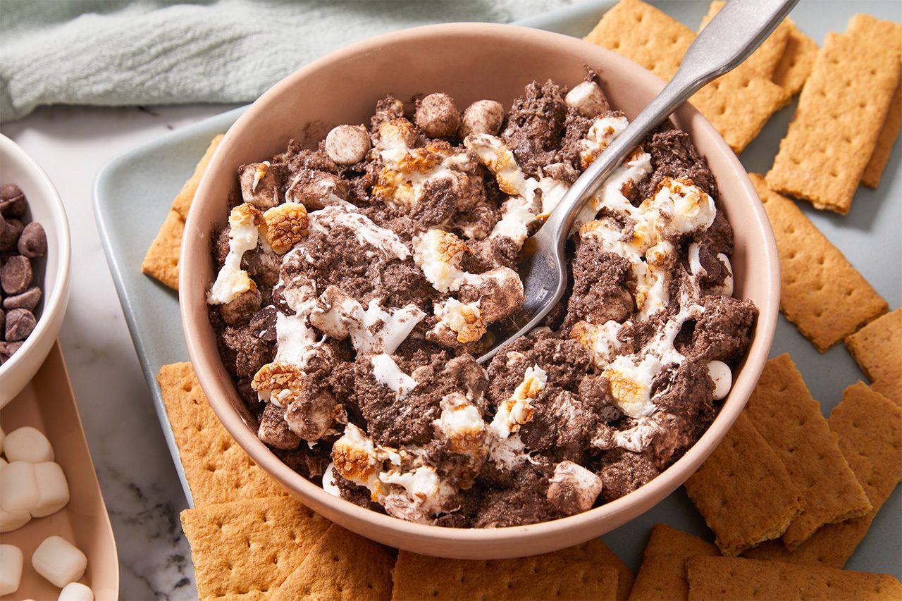 A bowl of s’mores cereal mixed with marshmallows and chocolate pieces, surrounded by graham crackers on a plate. A spoon is in the bowl, ready for eating.