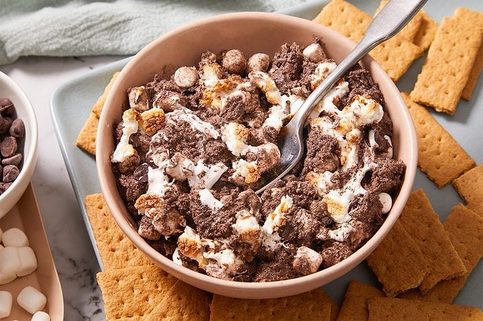 A bowl of s’mores cereal mixed with marshmallows and chocolate pieces, surrounded by graham crackers on a plate. A spoon is in the bowl, ready for eating.