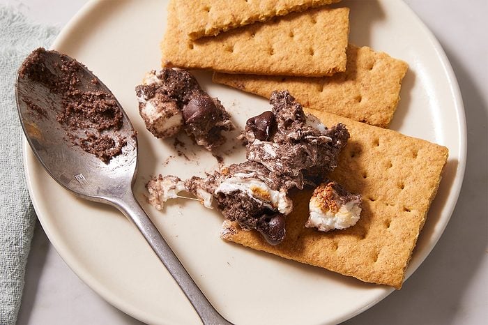 A plate with two rectangular graham crackers and a scoop of s’mores dip, featuring melted chocolate and toasted marshmallow, next to a metal spoon.