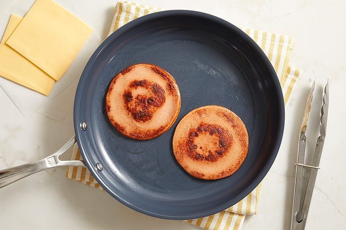 Two slices of fried bologna in a frying pan on a striped yellow towel, with a pair of metal tongs and slices of cheese nearby on a white surface.