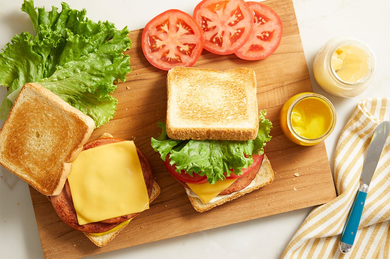 Two sandwiches with lettuce, tomato, cheese, and toast on a wooden board. Sliced tomatoes, leaf lettuce, a jar of mustard, and a knife on a napkin are also on the board.