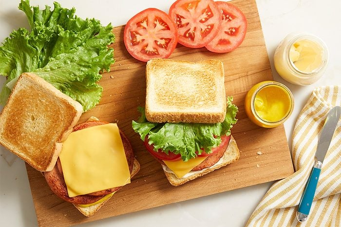 Two sandwiches with lettuce, tomato, cheese, and toast on a wooden board. Sliced tomatoes, leaf lettuce, a jar of mustard, and a knife on a napkin are also on the board.