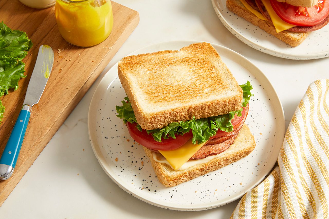 A plate with a toasted sandwich filled with lettuce, tomato, cheese, and deli meat. In the background are mustard, lettuce, and a knife on a cutting board, with a yellow-striped napkin nearby.