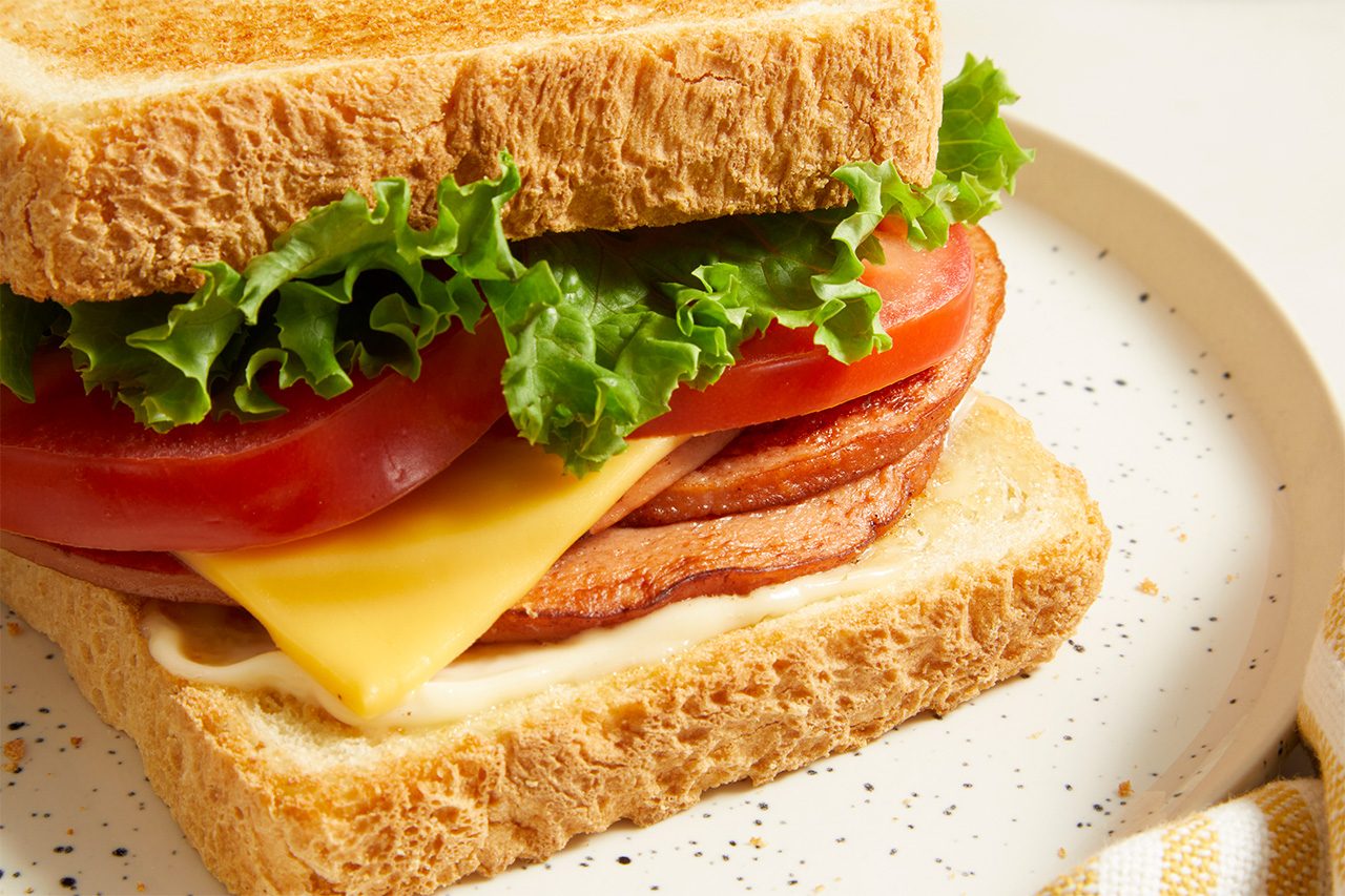 A close-up of a sandwich on a plate with whole wheat bread, lettuce, tomato slices, yellow cheese, and what appears to be slices of luncheon meat.