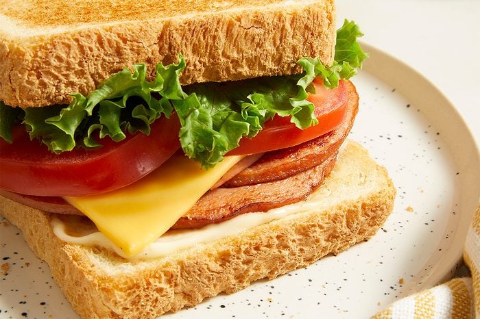 A close-up of a sandwich on a plate with whole wheat bread, lettuce, tomato slices, yellow cheese, and what appears to be slices of luncheon meat.