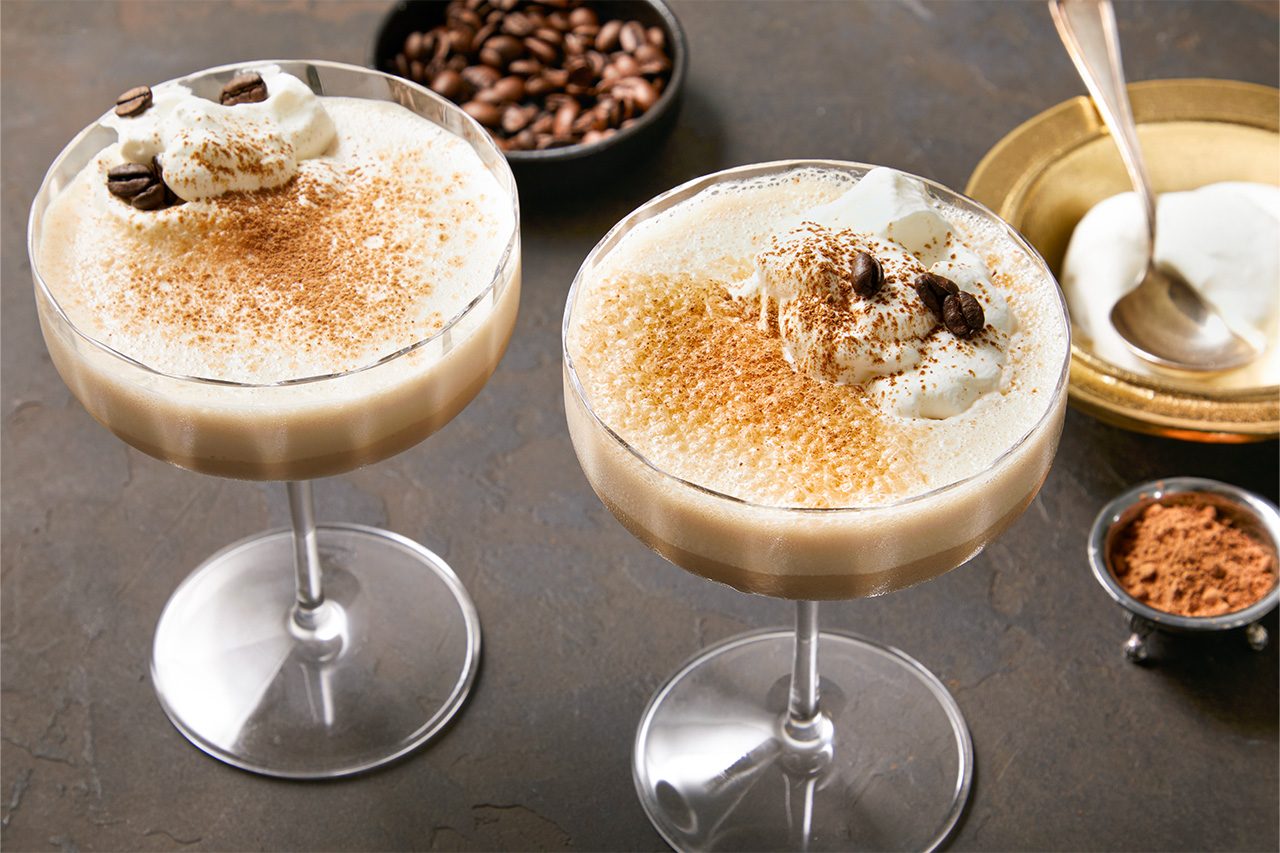 Eye Level horizontal image of two festive cocktails or creamy coffee drinks in stemmed glasses, garnished with whipped cream, cocoa powder, and whole coffee beans. A black bowl of coffee beans, a small gold dish with additional whipped cream, and a small silver bowl of cocoa powder are visible in the background.