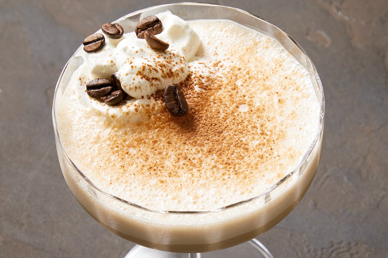 Overhead horizontal image of a creamy, frothy beverage served in a stemmed glass. The drink is generously topped with a dollop of whipped cream, a dusting of cinnamon powder, and garnished with several whole coffee beans. The light brown beverage has a rich, foamy texture, suggesting a coffee-based or dessert drink.