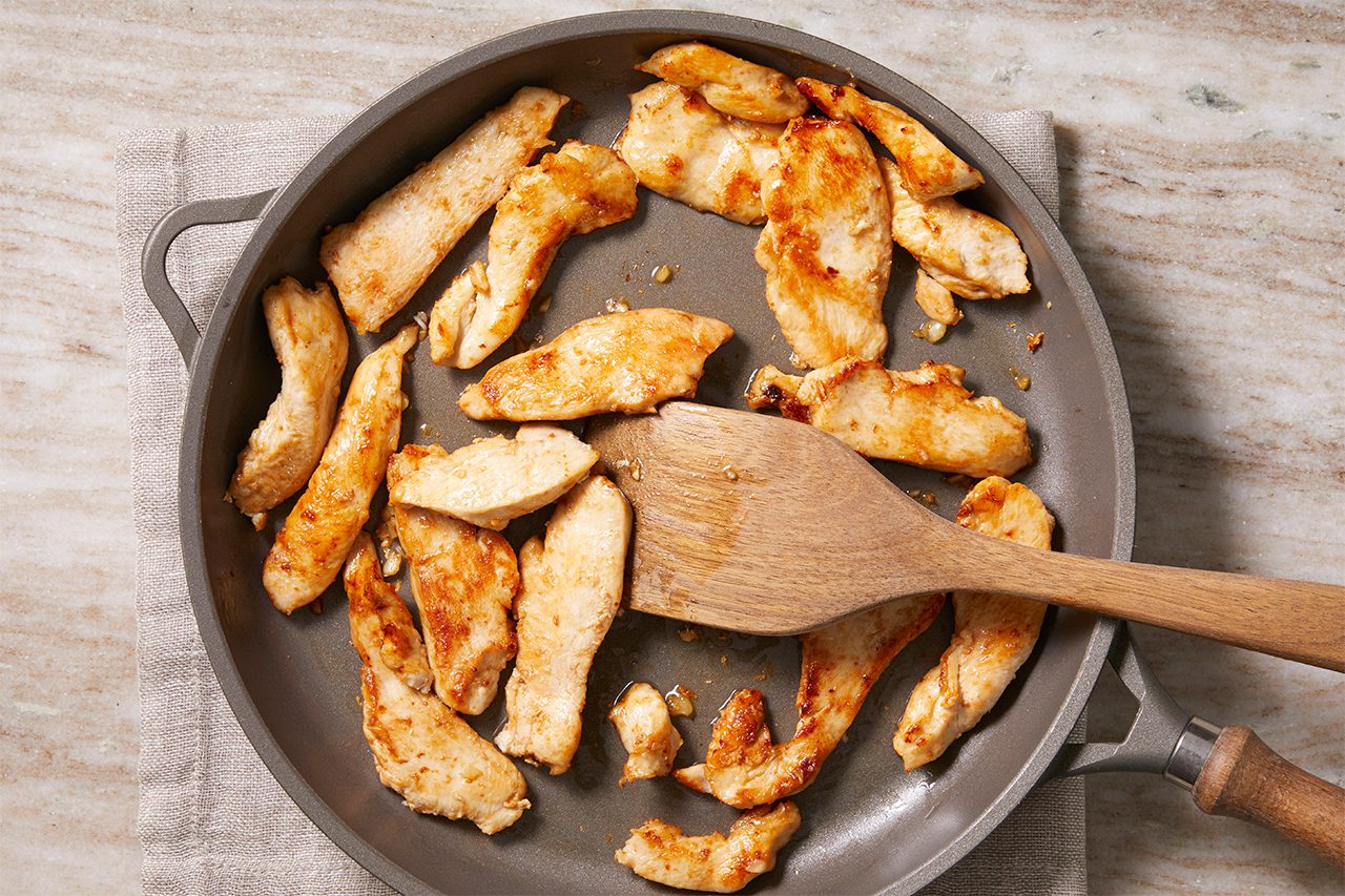 Overhead horizontal image of boneless skinless chicken breast strips browning in a pan with a wooden spatula. The cooked chicken and melted butter are visible in the pan, actively cooking.