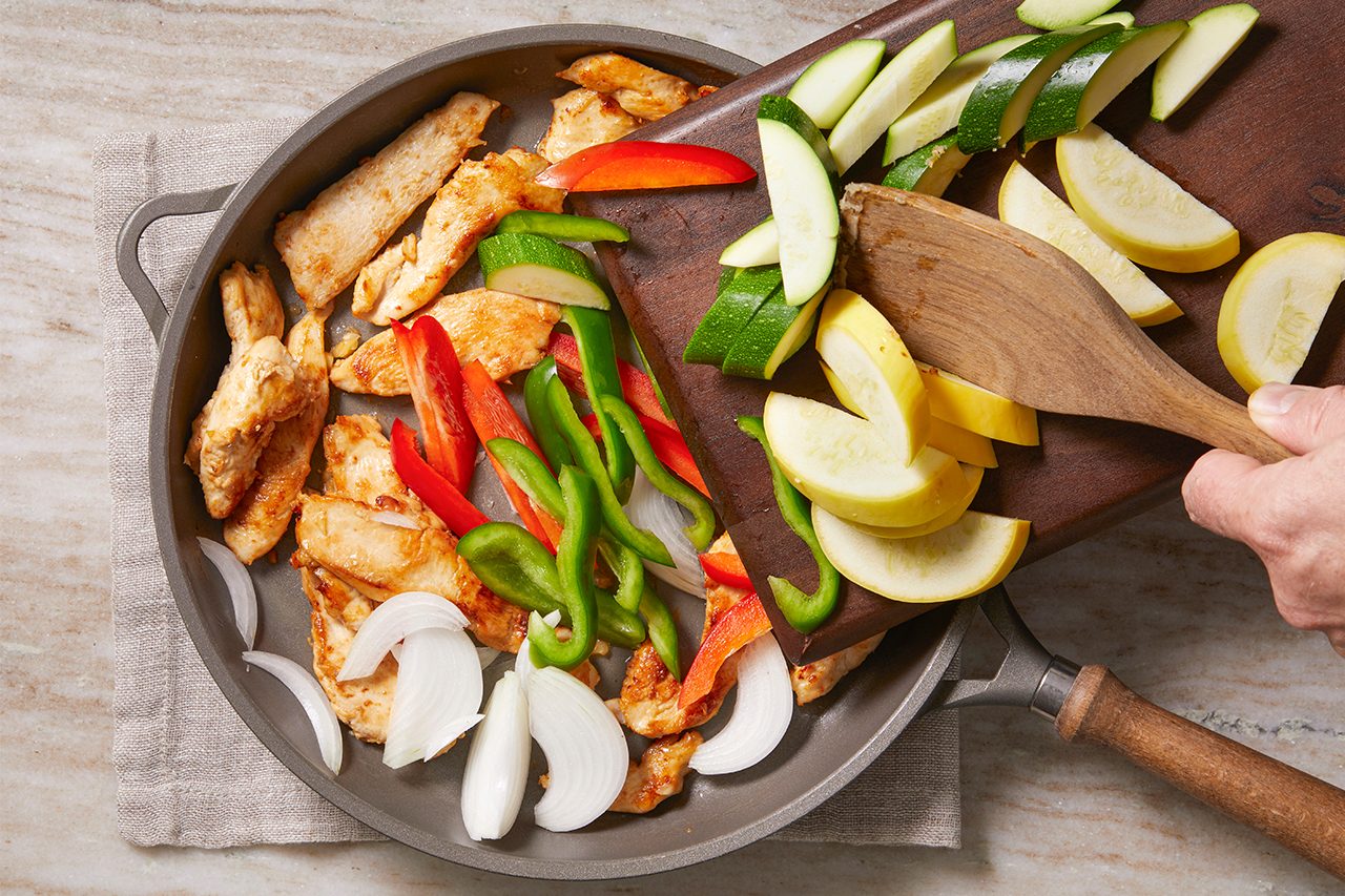 Overhead horizontal image of a hand sliding sliced yellow squash, zucchini, red pepper, and green pepper from a wooden cutting board into a skillet. The skillet contains cooked chicken strips and thinly sliced onions, preparing for the next stage of cooking.